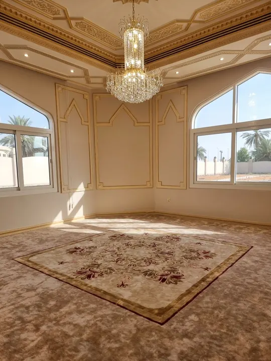 Salón vacío bajo una lámpara de cristal, con una alfombra floral con cenefa sobre moqueta